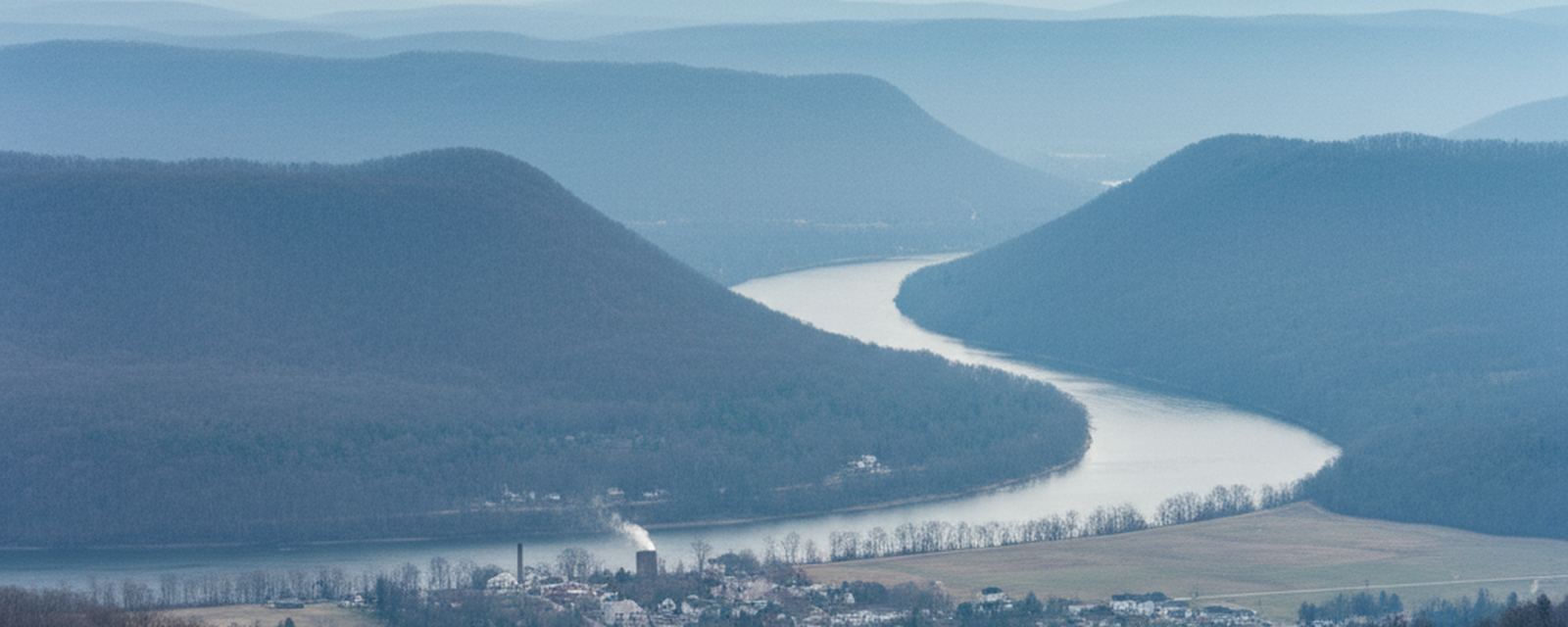 Hazy blue ridges layered above a wide river bend, and in the town below a tall smokestack and a second stack trailing a white plume stand among bare winter trees and an open field