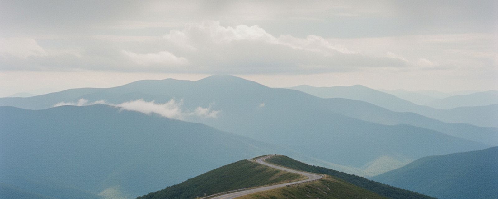 Two lane mountain road curving along a high ridge in the southern Appalachians, layered blue ranges falling away on both sides under low cloud.