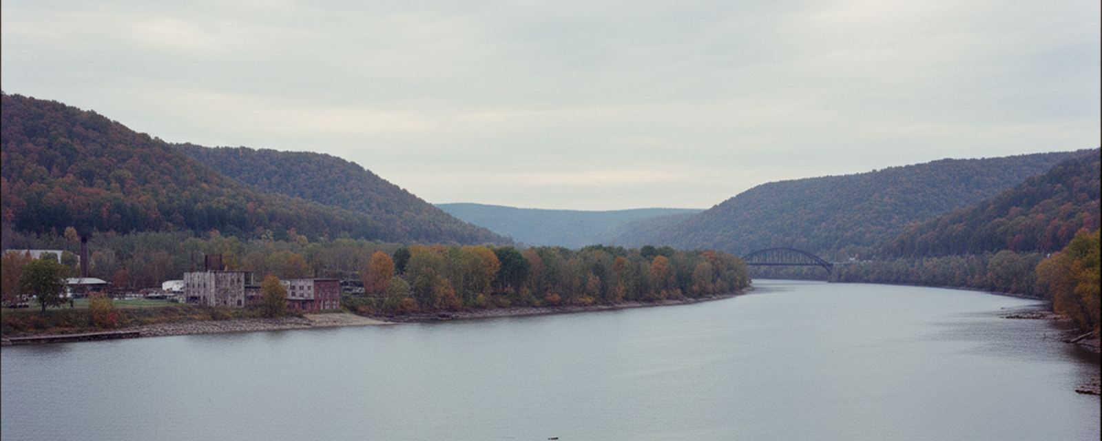 A wide river curves between wooded autumn hills, with an industrial plant and tall smokestack on the left bank and a steel arch bridge spanning the water in the distance.