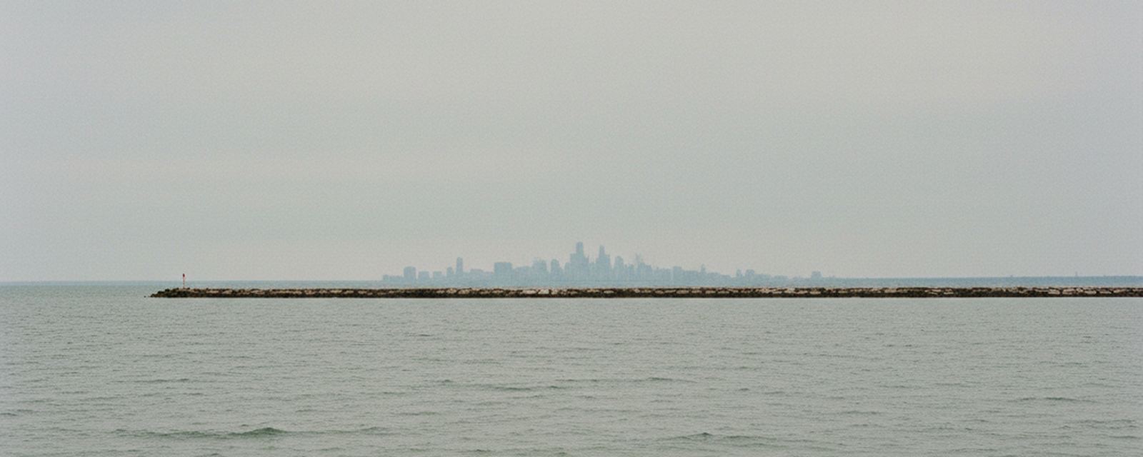Open gray water under flat overcast, a long rubble stone breakwater crossing the frame with a red and white marker post at its left end, and a hazy skyline of towers far behind it.