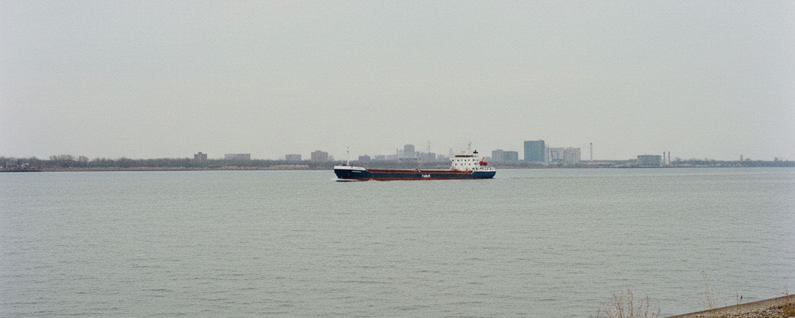The Detroit river from the paved near bank, wide gray water running toward Lake Erie, a low freighter in the channel and the far shore beyond it.