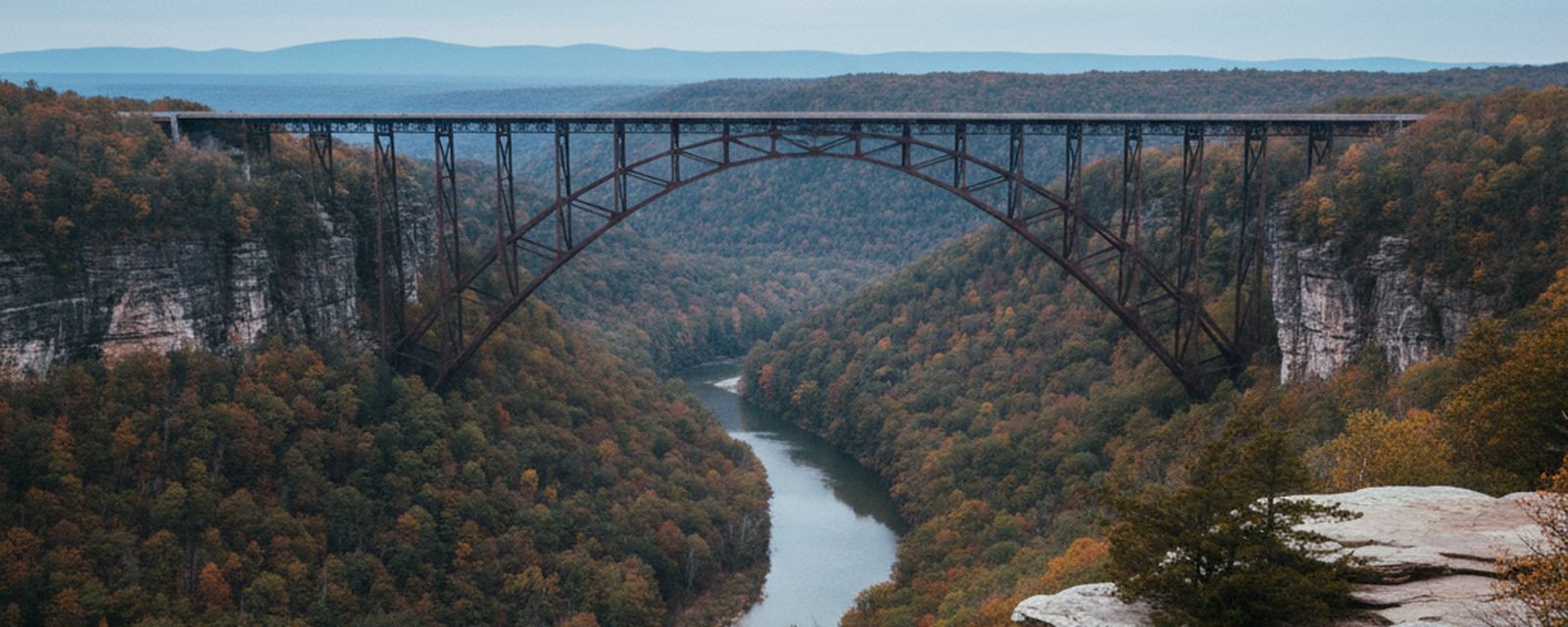 The steel arch over the New River seen from the north rim, sandstone ledges dropping into hardwood forest, the water narrow and pale far below.