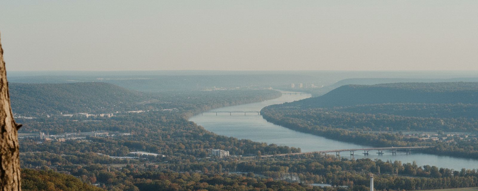 A wide river bends below a high wooded lookout, two bridges crossing it, a small town and low works on the bottomland, and pale ridges fading behind in autumn haze.